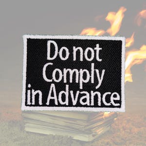 Könnte beinhalten: Ein schwarzer Patch mit weißem Text, der "Do not Comply in Advance" lautet. Der Patch liegt auf einem Stapel Bücher mit einem verschwommenen Bild von Flammen im Hintergrund.