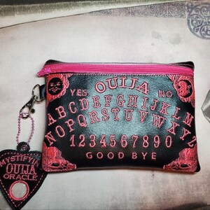 Puede incluir: Bolsa negra con cremallera con una cremallera rosa y un dise&ntilde;o de tablero de Ouija bordado en rosa. La bolsa tiene un peque&ntilde;o amuleto en forma de coraz&oacute;n rosa con las palabras "Mystify Ouija Oracle" en &eacute;l.