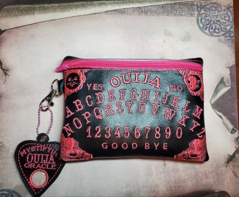 Puede incluir: Bolsa negra con cremallera con una cremallera rosa y un dise&ntilde;o de tablero de Ouija bordado en rosa. La bolsa tiene un peque&ntilde;o amuleto en forma de coraz&oacute;n rosa con las palabras "Mystify Ouija Oracle" en &eacute;l.