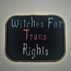 Op de afbeelding: Zwarte geborduurde patch met gouden stiksels. De tekst luidt "Witches For Trans Rights" in een gestileerd lettertype. Het woord "Trans" is roze, "Witches" is blauw en "Rights" is wit.