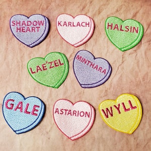 May include: Eight heart-shaped candy conversation hearts with various names from the video game Baldur's Gate 3. The names include: Shadowheart, Karlach, Halsin, Lae'zel,  Minthara, Gale, Astarion, and Wyll.