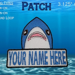 May include: Embroidered iron-on patch featuring a blue shark with a white mouth and teeth, holding a white banner with the text "YOUR NAME HERE". The patch is 3.125 inches by 3.5 inches.