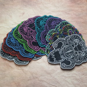 May include: A set of 10 embroidered iron-on patches featuring a black and white design of a creature with multiple eyes. The patches are layered in a rainbow of colours, including red, orange, yellow, green, blue, indigo, and violet.