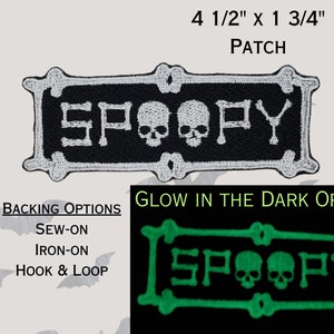Könnte beinhalten: Ein gestickter Patch in Schwarz und Weiß mit dem Wort "SPOOPY" in weißer Schrift und zwei weißen Totenköpfen. Der Patch ist 11,4 cm x 4,4 cm groß. Der Patch ist mit den Befestigungsmöglichkeiten zum Annähen, Aufbügeln und mit Klettverschluss erhältlich. Eine Version des Patches, die im Dunkeln leuchtet, ist ebenfalls erhältlich.