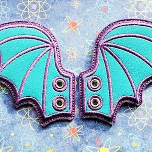 May include: A pair of teal blue fabric shoe laces with purple stitching and eyelets. The laces are shaped like bat wings.