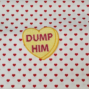 May include: A yellow heart-shaped patch with the words "DUMP HIM" embroidered in pink. The patch is set against a white background covered in small red hearts, creating a contrasting and playful design.