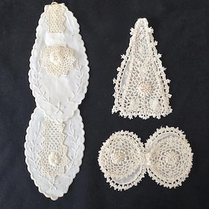 May include: A collection of white lace and embroidered items, including two oval-shaped placemats with floral and leaf designs, a triangular lace piece, and a double-circle lace piece, all against a dark background.