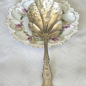 May include: Silver-toned serving utensil with a leaf-shaped head, featuring grape and leaf detailing. The handle has an ornate floral design. It rests on a floral patterned plate with a gold rim, set against a white background.