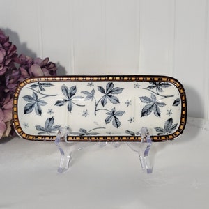 May include: A white ceramic soap dish with a brown and yellow border. The dish is decorated with a blue and white floral pattern. The dish is on a clear plastic stand.