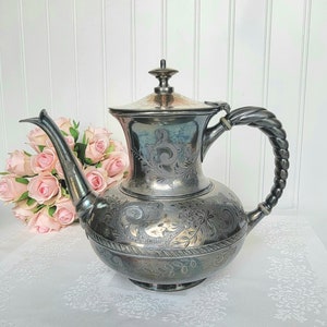 May include: A silver-colored antique teapot with intricate floral and scrollwork engravings. The teapot features a twisted handle and a spout. The lid is topped with a small finial. A bouquet of pink roses is in the background.