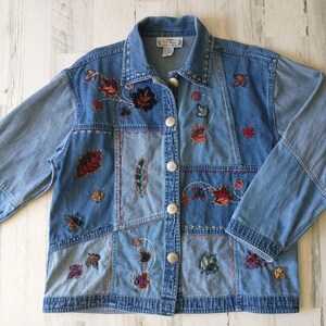 May include: A light blue denim jacket with a patchwork design. The jacket features embroidered leaves in shades of red, orange, and green. It has a button-up front and long sleeves. The jacket is a vintage style.