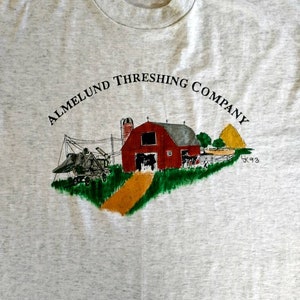 May include: A grey t-shirt with a vintage-style illustration of a red barn, a yellow path, and a farm scene. The text "ALMELUND THRESHING COMPANY" is printed in black above the illustration. The year "'93" is printed in black below the illustration.