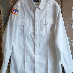 May include: White long-sleeve shirt with a button-down collar, two chest pockets, and a small American flag patch on the left sleeve. The shirt has a Blauer logo tag on the collar.