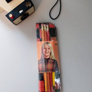 May include: A vintage pencil case with a black and red plaid design. It features a photo of a young woman with long blonde hair. The case contains four yellow pencils with red and blue accents.