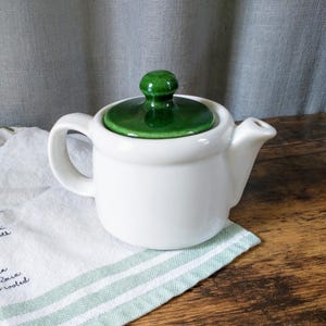 May include: A small, white ceramic teapot with a green lid and handle. The lid has a rounded green knob. The teapot is sitting on a white cloth with green stripes and text.