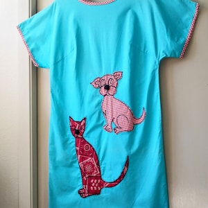 May include: A turquoise dress with short sleeves and a round neckline, trimmed with a red and white gingham pattern. The dress features appliqued designs of a red cat and a pink dog.