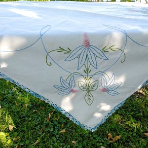 May include: White tablecloth with blue and pink embroidered floral designs. The tablecloth has a crocheted edge.
