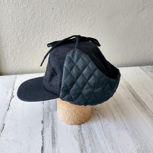 May include: A black, wool-blend hat with a quilted ear covering and a small brim. The hat is displayed on a cylindrical, woven stand. The ear covering is diamond-stitched. The hat has a drawstring closure.