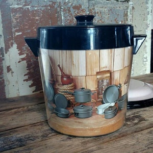 May include: A black and clear plastic beverage dispenser with a wood-grain design featuring a kitchen scene with various pots and pans. The dispenser has a lid and a handle.