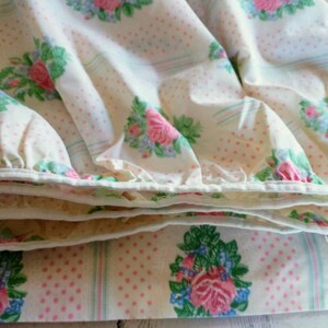 May include: Close-up of a folded floral bed sheet. The sheet features a pattern of pink roses, blue flowers, and green leaves on a cream background with pink polka dots and light blue stripes. The edges are white.