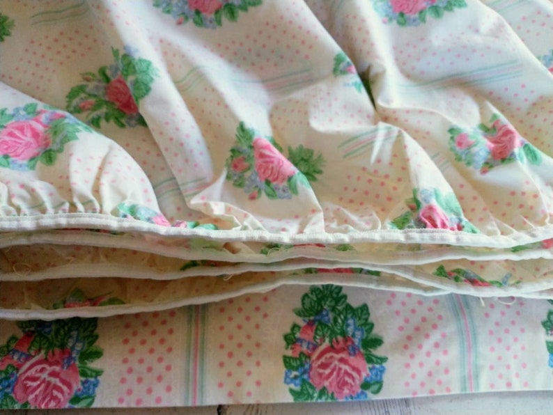 May include: Close-up of a folded floral bed sheet. The sheet features a pattern of pink roses, blue flowers, and green leaves on a cream background with pink polka dots and light blue stripes. The edges are white.