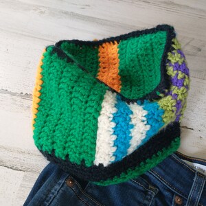 May include: A colorful crocheted neck warmer with stripes of green, orange, white, blue, and purple. The neck warmer is folded and lying on a pair of blue jeans.