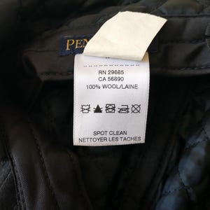 May include: Close-up of a black quilted fabric with a white tag. The tag reads "PEN" and includes details: RN 29685, CA 56890, and "100% WOOL/LAINE". The tag also has care symbols and the text "SPOT CLEAN".