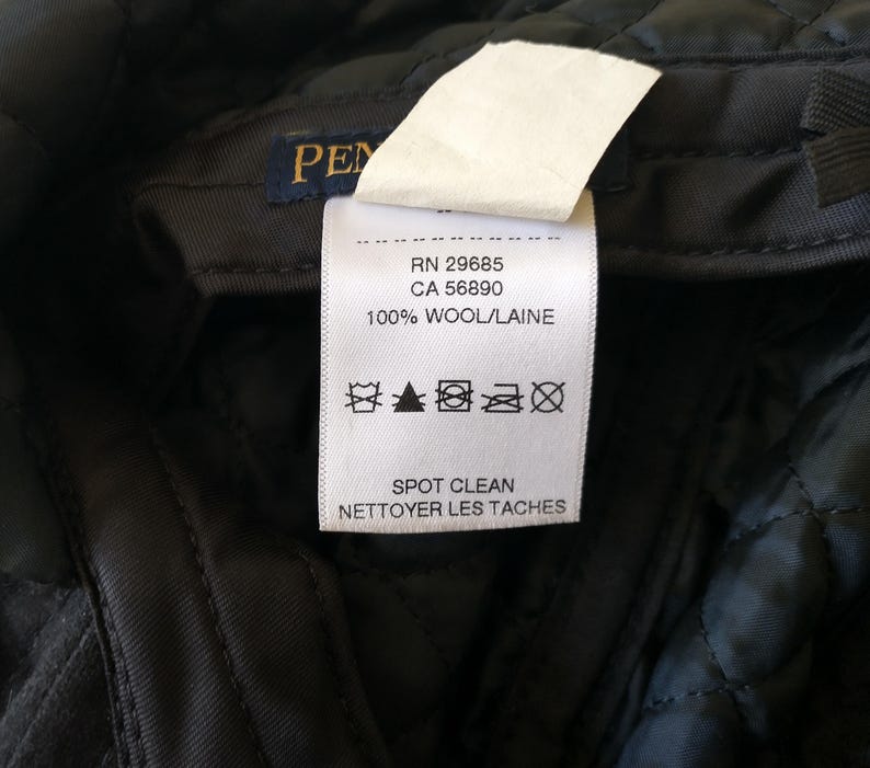 May include: Close-up of a black quilted fabric with a white tag. The tag reads "PEN" and includes details: RN 29685, CA 56890, and "100% WOOL/LAINE". The tag also has care symbols and the text "SPOT CLEAN".