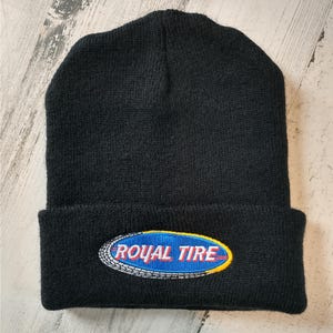 May include: Black knit beanie with a folded brim. The beanie features an embroidered patch with the words "ROYAL TIRE" in white, set against a blue oval with a yellow border. The patch is outlined with a tire tread design.