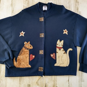 May include: Navy blue sweatshirt with a button-up front. The sweatshirt features appliqued designs of a dog, a cat, and stars. The dog and cat each have a heart. The buttons are square and have a plaid design.
