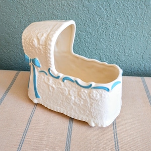 May include: White ceramic baby crib planter with a blue ribbon and blue trim. The planter is decorated with a lace-like pattern.