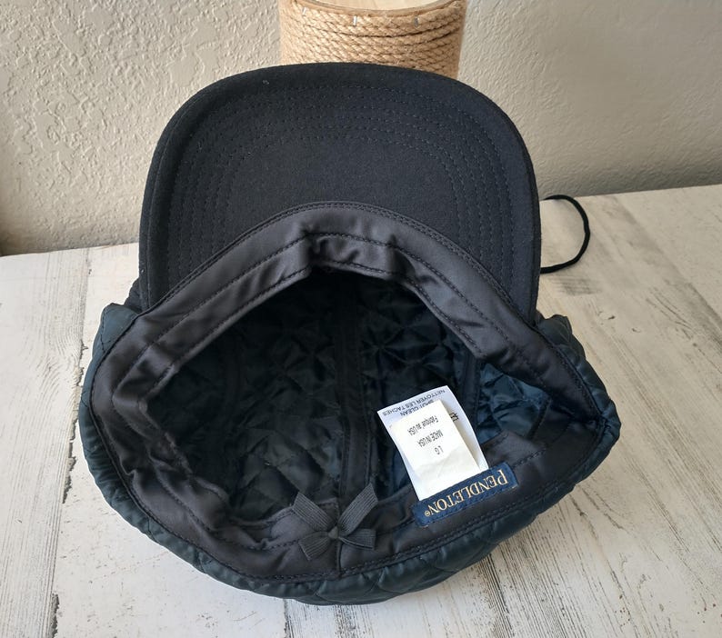May include: A black, quilted cap with ear flaps. The interior features a quilted lining and a tag that reads "PENDLETON". The cap is designed to provide warmth and protection from the elements.