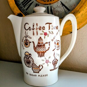 May include: White ceramic coffee pot with a lid and handle. The pot is decorated with brown text and illustrations, including the words "Coffee Time" and "No Sugar Please". A vintage clock is in the background.