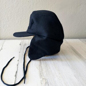 May include: A navy blue winter hat with a brim and ear flaps. The hat has a structured design and features long, black drawstrings for securing the ear flaps. The hat is made of a soft, dark fabric.