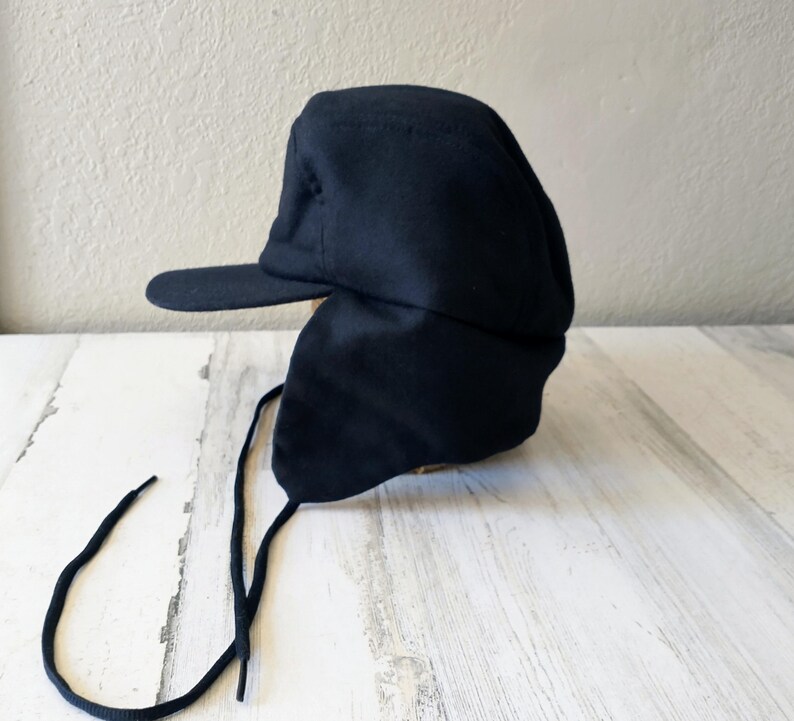 May include: A navy blue winter hat with a brim and ear flaps. The hat has a structured design and features long, black drawstrings for securing the ear flaps. The hat is made of a soft, dark fabric.