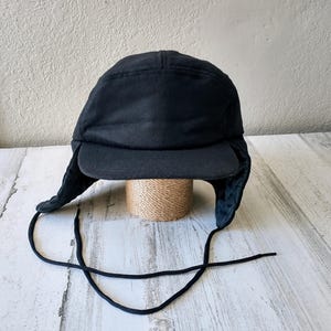 May include: A black winter hat with ear flaps and a brim. The hat is made of a dark, possibly wool, material. Black drawstrings hang from the ear flaps. The hat is sitting on a small, woven, cylindrical object.