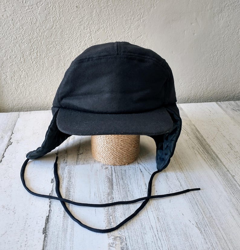 May include: A black winter hat with ear flaps and a brim. The hat is made of a dark, possibly wool, material. Black drawstrings hang from the ear flaps. The hat is sitting on a small, woven, cylindrical object.