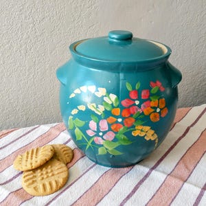 May include: Teal ceramic cookie jar with a lid, adorned with hand-painted flowers in shades of pink, red, orange, and yellow. The jar sits on a striped cloth next to three peanut butter cookies.
