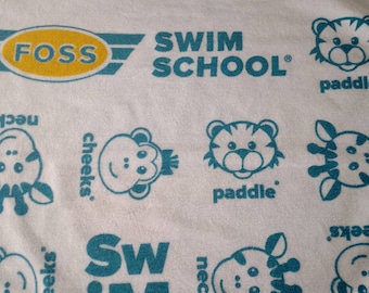 Könnte beinhalten: Weißes Handtuch mit einem blauen und gelben Muster. Das Muster enthält die Wörter "Swim School", "Foss" und "paddle". Es gibt auch Bilder von Affen, Giraffen und Tigern.