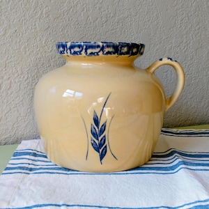May include: A yellow ceramic pitcher with a blue wheat stalk design. The pitcher has a rounded body, a handle, and a decorative blue rim. It sits on a blue and white striped cloth.