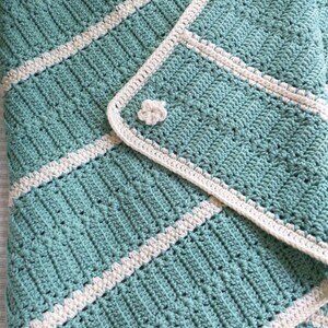May include: A handmade crochet blanket in shades of teal and cream. The blanket features a textured stitch pattern with diagonal stripes and a flower-shaped button closure. The soft, cozy blanket is perfect for a baby or as a decorative throw.