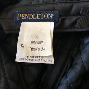May include: Close-up of a dark gray quilted fabric with a tag. The tag reads "PENDLETON&reg;" in gold lettering. Below the brand name, the tag indicates "LG", "MADE IN USA", and "SPOT CLEAN".