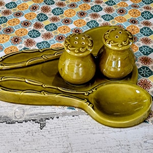 May include: A green ceramic salt and pepper shaker set with a matching spoon rest. The set features a unique design with a spoon-shaped base and a raised rim.