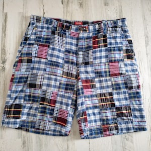 May include: Blue plaid shorts with a patchwork design. The shorts feature a mix of blue, red, black, and white plaid patterns. The brand name "IZOD" is visible on the waistband. These shorts are a casual summer garment.