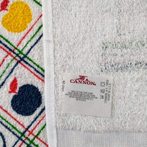 K&ouml;nnte beinhalten: Wei&szlig;es Baumwollhandtuch mit einem geometrischen Muster in Rot, Gr&uuml;n, Blau und Gelb. Das Handtuch hat einen Fransenrand und ein wei&szlig;es Etikett mit dem Text "CANNON" und Pflegehinweisen.