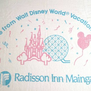 May include: A white towel with a pink flamingo, a blue and pink castle, a blue and white sphere, a pink balloon with Mickey Mouse ears, and a palm tree. The text reads "One mile from Walt Disney World® vacation Kingdom Radisson Inn Maingate".