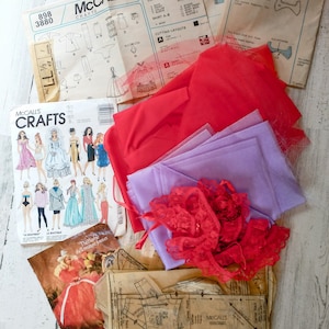 May include: A collection of vintage sewing patterns and fabric scraps for a doll's nightgown and robe. The patterns are from McCall's Crafts and include instructions for a top, coat, skirt, dress, and scarf. The fabric scraps are red, purple, and white.