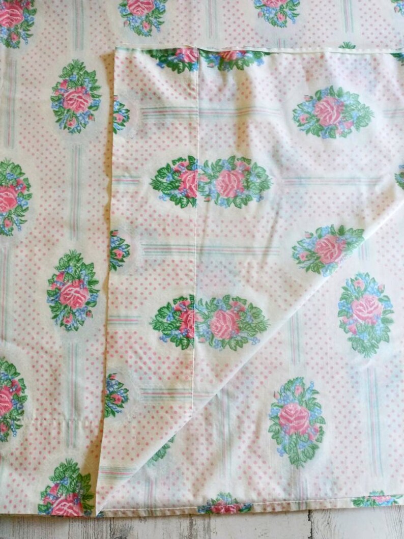 May include: A folded white sheet with a repeating pattern of pink roses, blue accents, and green leaves. The design is set against a backdrop of small pink polka dots and vertical stripes.