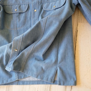 May include: A light blue denim shirt featuring a button-down front with silver-toned metal snap closures, two chest pockets with flaps, long sleeves, and side slits at the hem. The fabric has a visible vertical weave pattern and light blue stitching.
