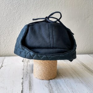 May include: A dark navy blue winter hat with a quilted brim and ear flaps. The hat has a soft, felt-like crown and a tie closure at the top. The hat is displayed on a cylindrical stand wrapped in natural twine.